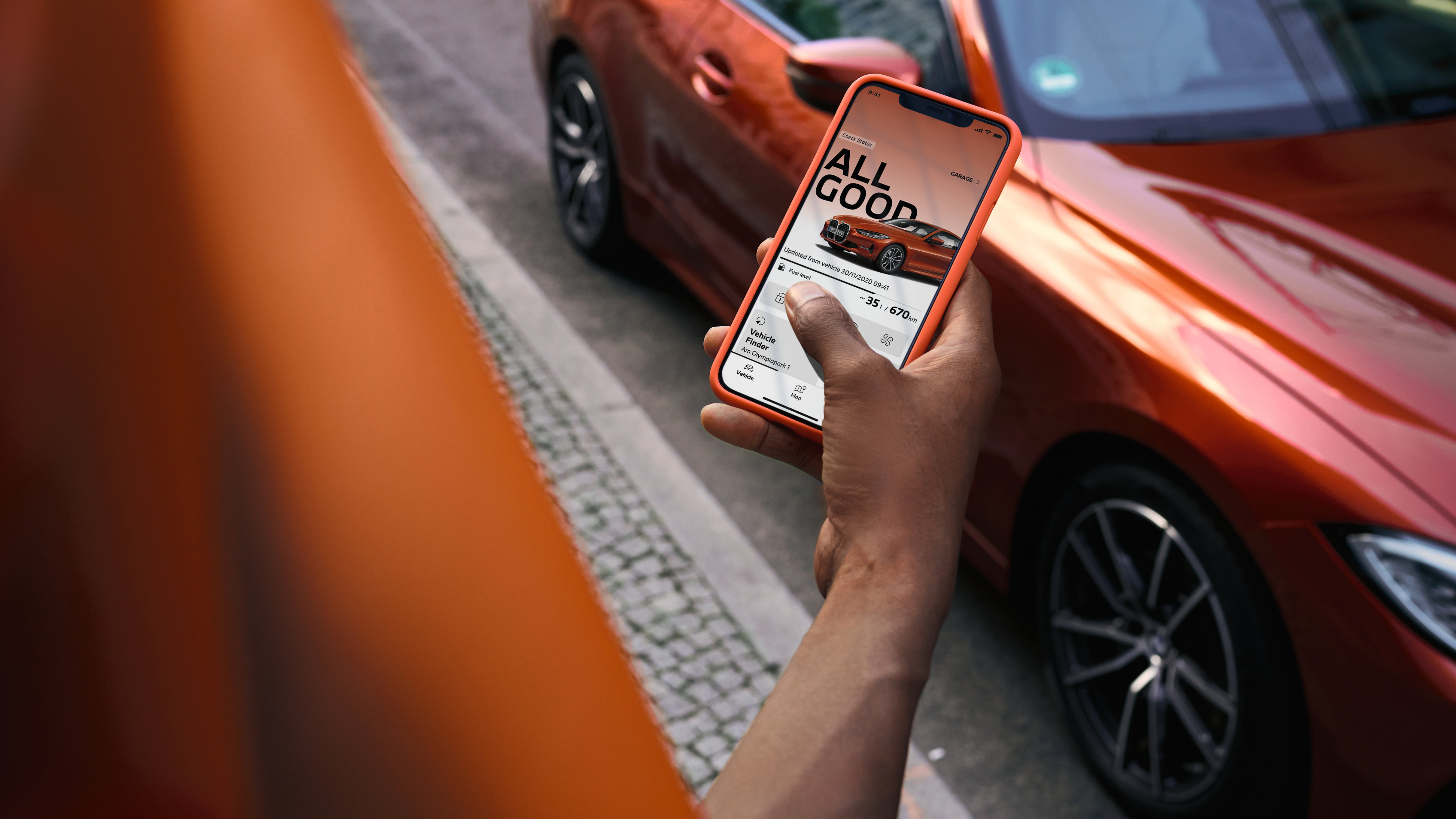A hand holds a smartphone showing the MyBMW App on screen. An orange BMW sits in the background.