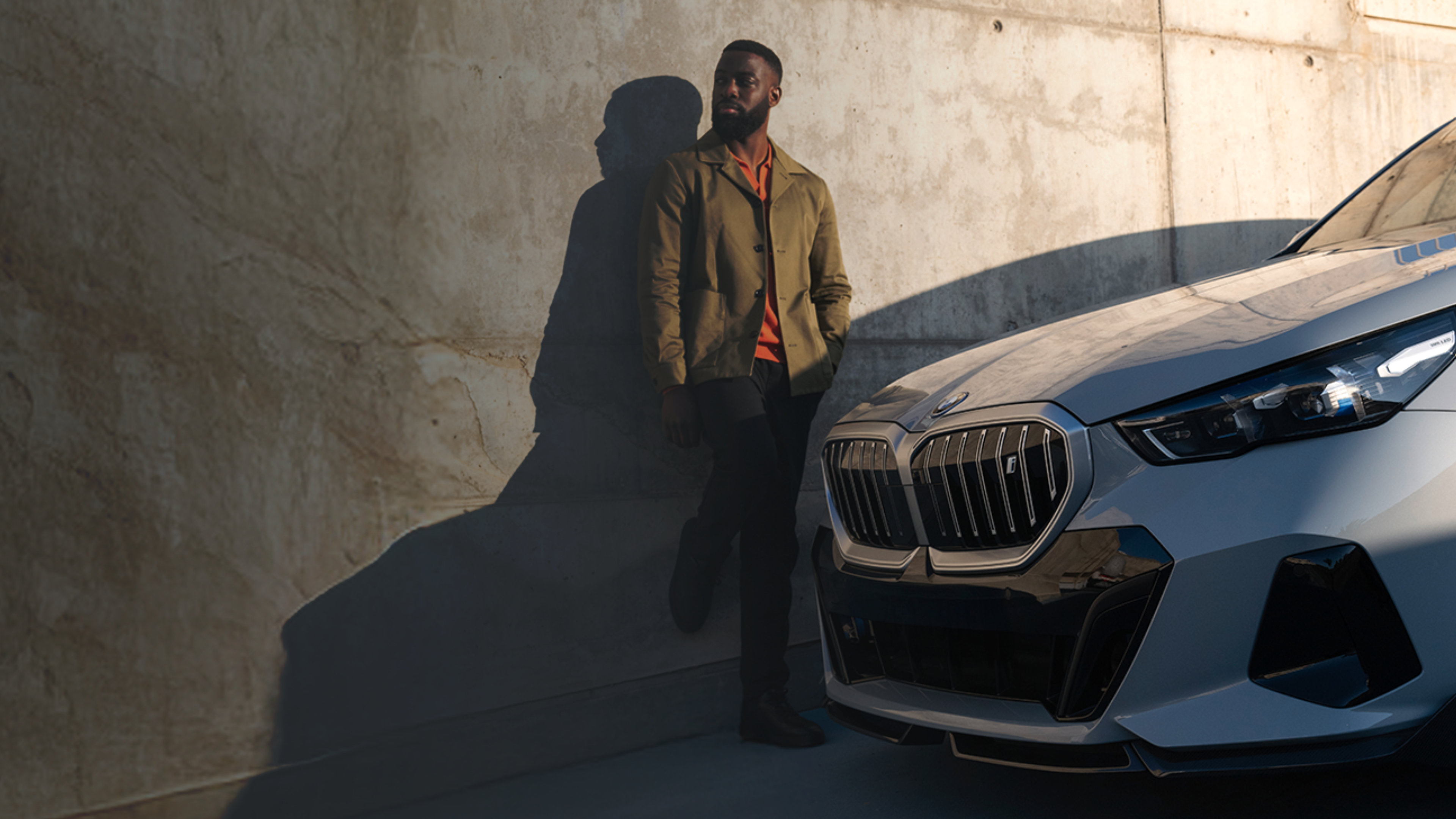 A silver BMW i5 eDrive40 is parked next to a wall. A man in a tan coat and orange shirt leans on the wall next to the car.