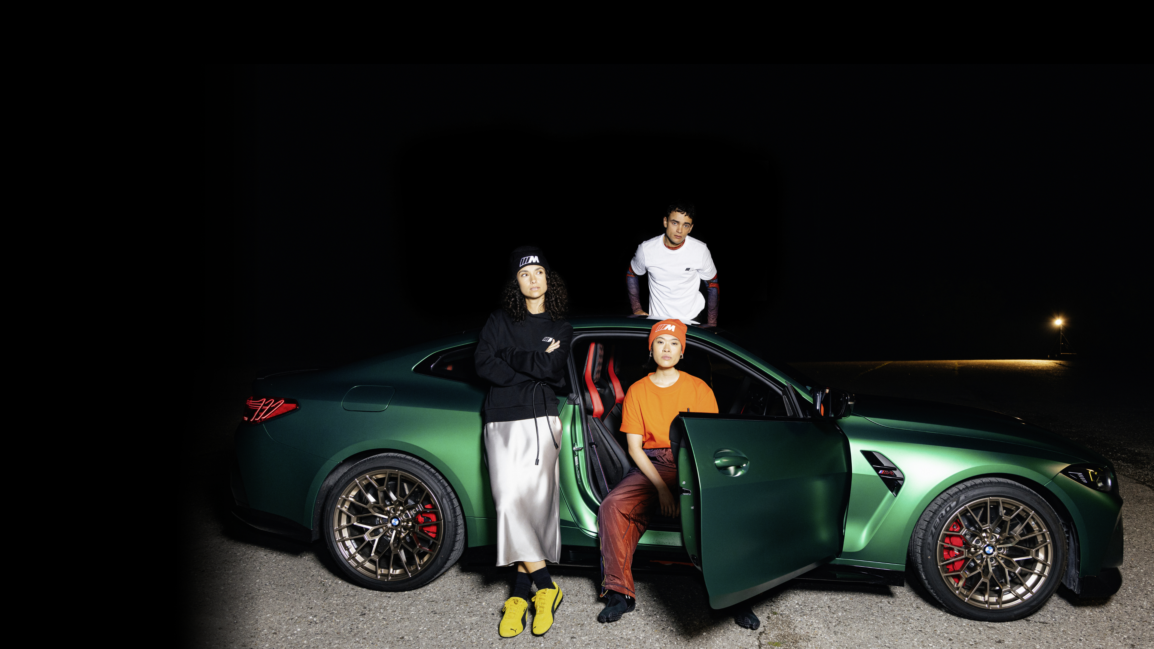 Two women and a man wearing M-branded apparel stand by a green BMW M5