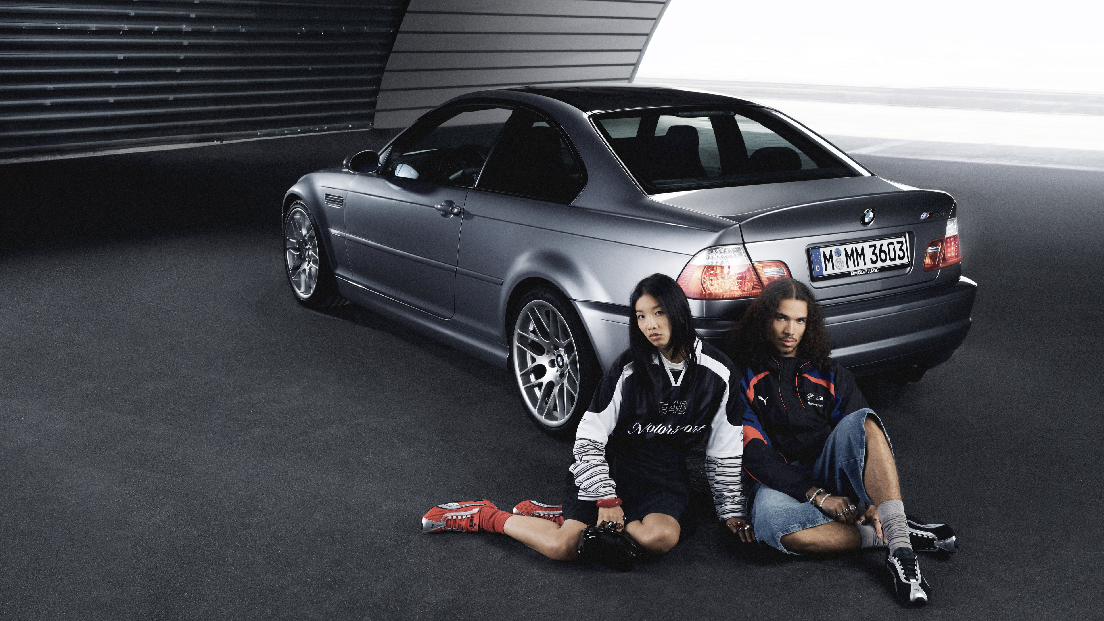 A young woman and young man is BMW Motorsport apparel sit behind the rear bumper of a European model BMW coupe.