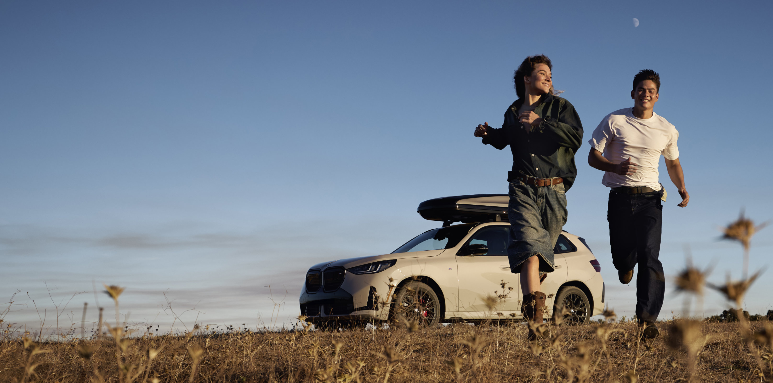 A white BMW X3 with black wheels and a BMW Roof Box installed on its roof is parked in a field. A man and a woman run across the field towards the camera.