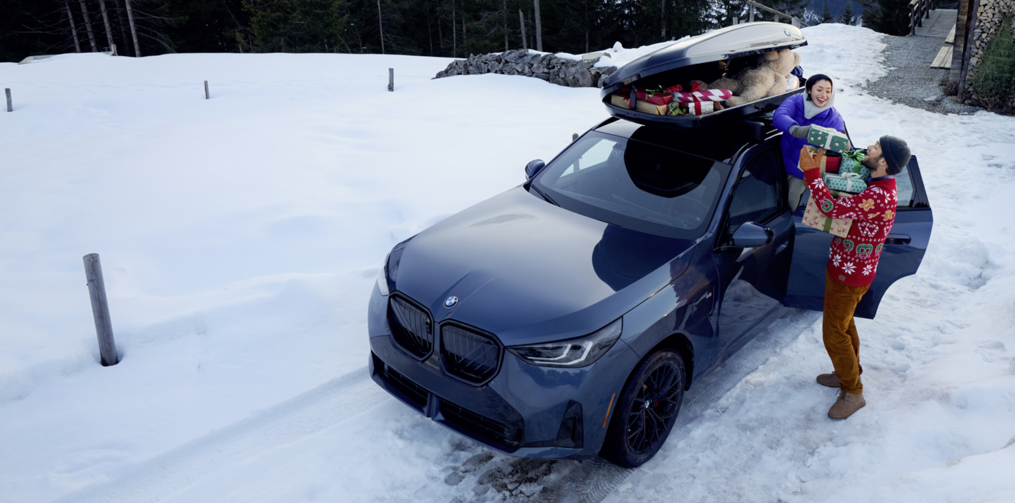 A blue BMW X3 with a BMW roof box full of gifts and toys installed is parked in a snowy mountainside driveway.